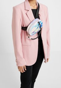 Holograficzna torebka crossbody z zamkiem błyskawicznym i czarnym łańcuchowym paskiem, noszona na różowej marynarce i czarnym topie, o zaokrąglonym kształcie.
