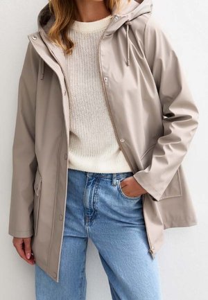 Kobieta w beżowej kurtce przeciwdeszczowej z kapturem, na białym dzianinowym swetrze i jasnoniebieskich dżinsach, z jedną ręką w kieszeni.