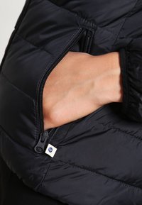 Černá prošívaná bunda s bočním kapsou na zip, hladkým nylonovým povrchem, prošívanou strukturou a malou logovou cedulkou u otevření kapsy.