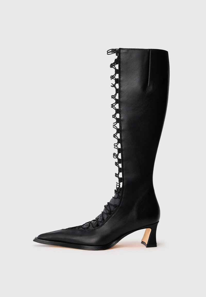 Μαύρη δερμάτινη μπότες μέχρι το γόνατο με μυτερή μύτη, κορδόνια μπροστά και μικρό χοντρό τακούνι. Διαθέτει υφή με ανάγλυφα σχέδια στις πλευρές.