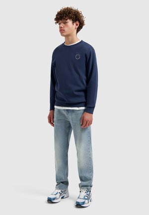 Jonge man die een marineblauw ronde hals sweatshirt draagt, lichtblauwe jeans en witte sneakers met blauwe accenten, staande tegen een effen lichte achtergrond.