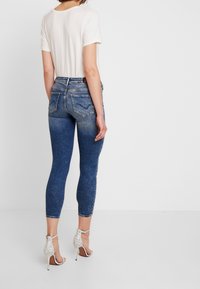 Vaqueros ajustados en denim azul oscuro con una cintura de media altura, que cuentan con bolsillos traseros y un ligero desvanecimiento. Combinados con tacones abiertos blancos.