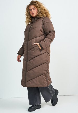 Femme aux cheveux bouclés portant un long manteau matelassé marron, un pantalon noir à jambes larges et des bottes noires épaisses, marchant sur un fond uni.