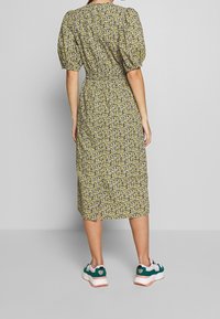 Midi šaty s nafouknutými rukávy v žlutě květinovém potisku, s úzkým pasem, po kolena a se slits na zádech, kombinované s robustními tyrkysovými a bílými teniskami.