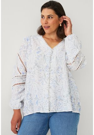 Vrouw draagt een witte blouse met bloemmotief en kanten inzetstukken op de mouwen en een blauwe spijkerbroek, raakt haar haar aan en kijkt met een lichte glimlach opzij.