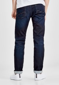 Jeans de denim oscuro con un corte ajustado, que presentan bolsillos traseros con costura curved, puños enrollados y una textura suave. Combinados con zapatos blancos.