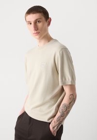 Beige, rövid ujjú póló, puha anyagból készült, kicsi logós jelvényt tartalmaz a mellrészen és laza szabású.