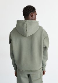 Sweat à capuche en vert olive, fabriqué en matériau doux. Il présente une coupe décontractée, des coutures visibles et une poche kangourou.