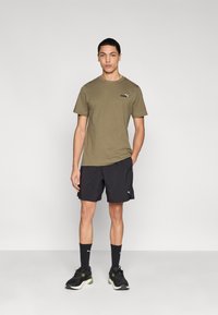 Χακί βαμβακερό μπλουζάκι με μικρό μαύρο λογότυπο, συνδυασμένο με μαύρο σορτς, μαύρες κάλτσες και μαύρα αθλητικά παπούτσια με πράσινες λεπτομέρειες.