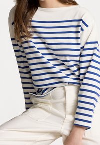 Woman wearing a white and blue striped long-sleeve shirt and off-white high-waisted pants, seated with one knee raised.