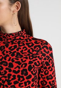 Top de cuello vuelto rojo con patrón de leopardo negro. Presenta una textura suave. Accesorio: pendientes dorados que parecen alfileres de seguridad.