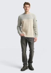 Junger Mann steht mit einer Hand in der Tasche, trägt einen zweifarbigen grauen und beigen Strickpullover, ausgebleichte schwarze Jeans und dunkle Schnürstiefel.