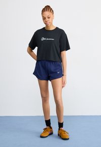 Črn krojeni majica s kratkimi rokavi z logotipom, mornarsko modri rebrasti šortsi in rjavi športni čevlji s vijoličnimi detajli. Preprost dizajn, sproščen kroj in lahek material.