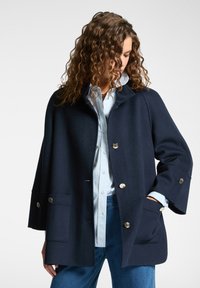 Cappotto in misto lana blu navy con una forma rilassata, dotato di colletto, due ampi taschini frontali e bottoni in tonalità argento. Texture liscia con un design strutturato.