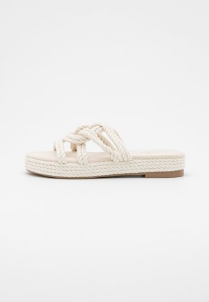 Beige flad sandal med tykke flettede reb stropper og en flettet platformsål på en hvid baggrund.