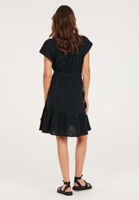 Zwarte, knielange jurk met korte mouwen, een ceintuur in de taille en een gelaagde rucheszoom. Gemaakt van lichtgewicht stof met een gestructureerde afwerking.