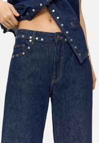 Dark blue denim jeans with silver metal accents along the pockets and button details. Features a straight cut and visible stitching.