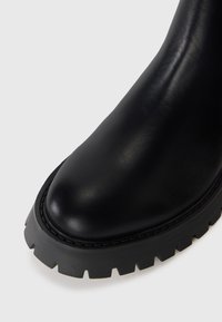 Botte en cuir noir avec un bout arrondi et une semelle à crampons épaisse, présentée sur un fond blanc.