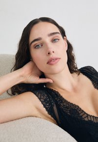 Zwart kanten kledingstuk met een delicate, golvende halslijn. De stof heeft een doorschijnende kwaliteit en toont ingewikkelde bloemmotieven. Zachte, gestructureerde achtergrond.
