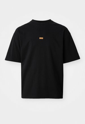Black cotton t-shirt with a relaxed fit, short sleeves, round neckline, and a tan logo patch on the chest. Smooth texture, minimal design.