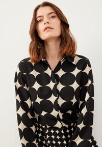 Zwarte en beige blouse met een groot cirkelpatroon, lange mouwen en een kraag, gemaakt van een gladde stof met een lichte glans.