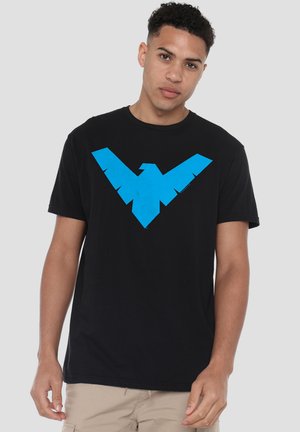 Junger Mann mit kurzen lockigen Haaren, der ein schwarzes T-Shirt mit einem großen blauen stilisierten Vogel-Logo auf der Brust und beige Hose trägt.