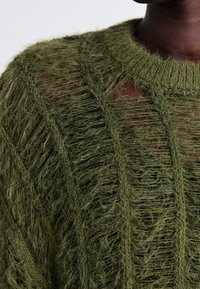 Grüner Strickpullover mit loser, strukturiertem Gewebe. Verfügt über offene Bereiche und vertikale Linien, mit einem gerippten Halsauschnitt. Weiches, leichtes Material.