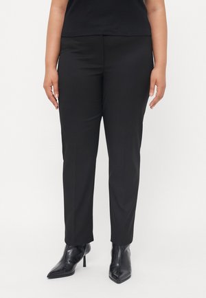 Femme portant un pantalon noir cintré et des bottes noires à talons hauts et bouts pointus, debout sur un fond blanc uni.
