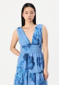 Gaiši zils ziedu pārklājs ar dziļu V veida kakla izgriezumu un platām siksnām. Krūšu daļai ir drapējums, un slāņotais svārku dizains ir plūstošs.