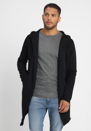 Hoodie negro largo hecho de tela suave con cremallera frontal completa, que presenta una capucha, bolsillos laterales y un corte relajado sobre una camiseta gris y jeans azules.