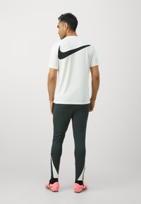 Polo shirt blanc Nike avec logo noir, associé à un pantalon côtelé vert foncé avec des accents blancs et des chaussures de sport roses.
