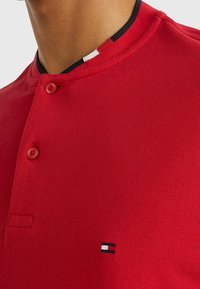 Röd polotröja med texturerat tyg, två knappar i knappslån och en liten logotyp i marinblått, vitt och rött nära nederkant.