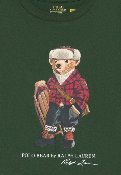Tricou verde cu o ilustrație de urs îmbrăcat într-o jachetă carouri roșie, pălărie gri și ținând un sanie, purtând pantaloni și cizme negre.