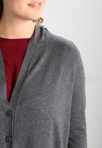 Szary kardigan z dekoltem w szpic i trzema guzikami, wykonany z miękkiej wełny, o gładkiej strukturze i dopasowanej sylwetce, noszony na burgundowej bluzce.