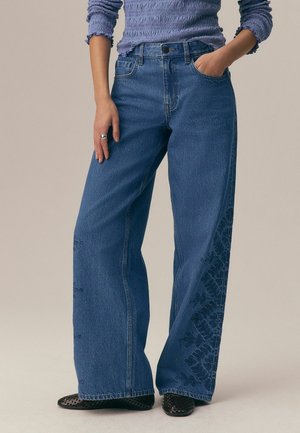 Žena nosí modré rifle s rozšírenými nohavicami a kvetinovou výšivkou na vonkajších stranách nôh, fialový textúrovaný top s dlhými rukávmi, čierne tkané topánky a prsteň.