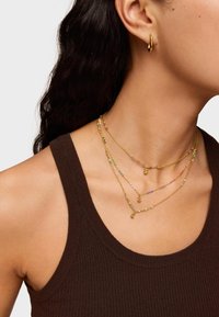 Collares en capas con cadenas de oro y cuentas de colores. Un pendiente de aro dorado suave complementa el look sobre una camiseta de tirantes oscura.