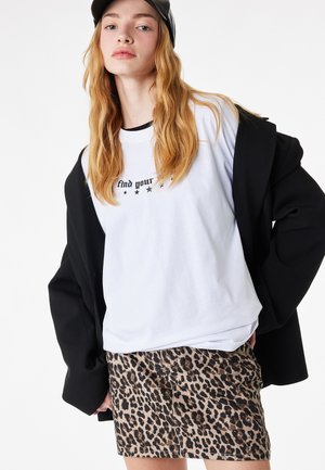 Ung kvinde med langt hår iført sort kasket, oversized sort jakke, hvid t-shirt med grafik og leopardmønstret nederdel mod hvid baggrund.