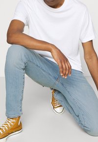 T-shirt blanc à manches courtes associé à un jean bleu clair et des baskets montantes marron. La tenue présente un style décontracté et simple.