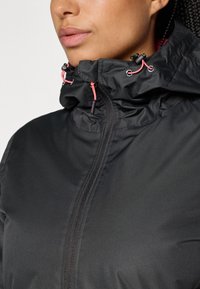 Veste noire imperméable avec un col haut, présentant un tissu froncé, des cordons de serrage rose et une fermeture éclair intégrale. Texture lisse.