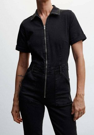 Nainen pukeutuneena istuvaan mustaan lyhythihaiseen haalariin, jossa on etuvetoketju, kaulus ja käärittyjen hihojen reunat, yksiväristä taustaa vasten.