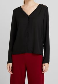 Blusa nera a maniche lunghe con scollo a V, caratterizzata da una chiusura con bottoni e dettagli in pizzo sulle spalle, abbinata a pantaloni bordeaux.