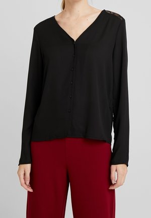 Blusa nera a maniche lunghe con scollo a V, caratterizzata da una chiusura con bottoni e dettagli in pizzo sulle spalle, abbinata a pantaloni bordeaux.