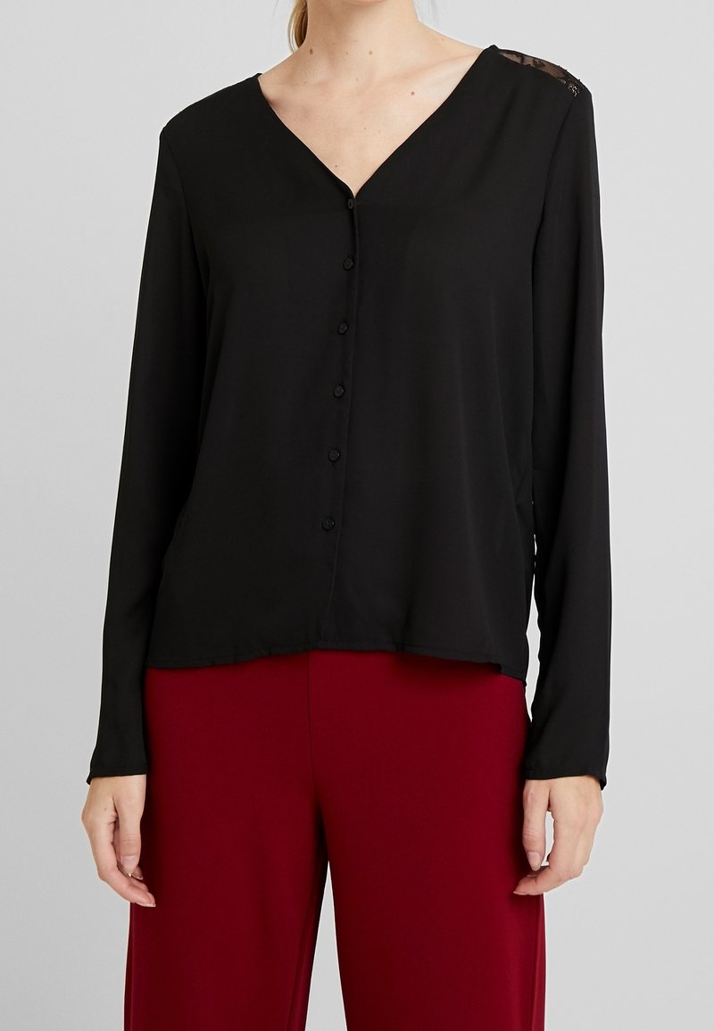 Blusa nera a maniche lunghe con scollo a V, caratterizzata da una chiusura con bottoni e dettagli in pizzo sulle spalle, abbinata a pantaloni bordeaux.