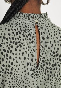 Blouse verte à motifs avec un col à volants et un dos ouvert fermé par un bouton. Des taches noires créent un design contrastant.