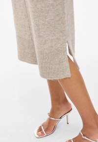 Bézs színű kötött pulóver bordázott szegéllyel és oldalsó hasítékokkal, ezüst színű pántos magassarkú szandállal párosítva. Puha tapintású, kényelmes szabás.