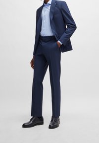 Traje azul marino con corte entallado, tejido texturizado, chaqueta de un botón y pantalones de frente plano. Camisa azul claro y zapatos de cuero negro.