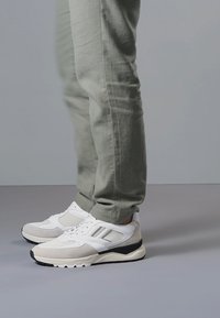 Osoba nosząca jasnoszare spodnie i białe buty sportowe z czarnymi podeszwami, stojąca na szarej podłodze na tle szarej ściany.