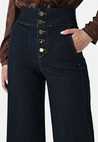 Pantalons en denim taille haute de couleur bleu foncé avec des boutons dorés, coutures contrastantes et poches latérales, présentant une coupe ajustée et un matériau robuste.