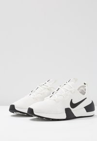 Valkoiset urheilujalkineet, joissa on teksturoitu verkkomateriaali, mustat yksityiskohdat ja kumipohja. Ominaisuuksiin kuuluvat nauhat ja brändin logo takanapissa.