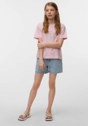 Young girl with long hair wearing a pink starfish T-shirt, light denim shorts, and silver sandals, standing with arms crossed on white background.